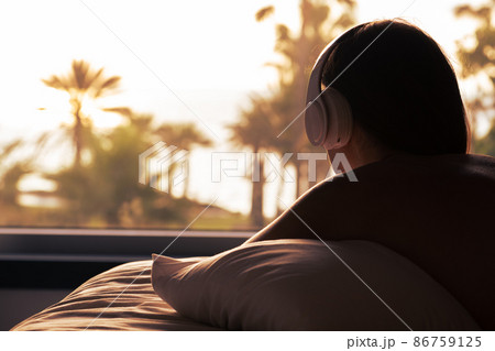 Silhouette of woman with headphones lying on pillow on the bed in bedroom opposite panoramic window with view on palm tree sea beach at sunset. Female relaxing listening music. 86759125