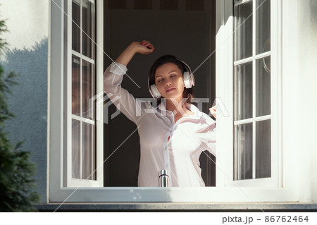 Young beautiful happy woman in headphones listening music opens window of country house and breathes fresh air in the early summer morning. Young beautiful happy woman in headphones listening music opens window of country house and breathes fresh air in the early summer morning. 86762464