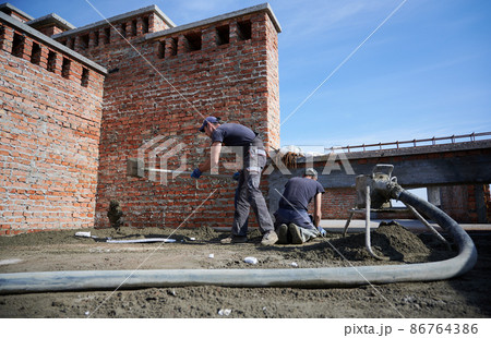 Two workers in process of applying cement mixture supplied by mobile mortar pump on work surface. Installation of semi-dry floor screed outside under open blue sky. 86764386