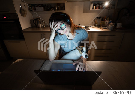 Tired woman in headphones and glasses studying at a laptop. A female student prepares for an exam in the kitchen late in the evening. Girl with a puppy on her knees. Tired woman in headphones and glasses studying at a laptop. A female student prepares for an exam in the kitchen late in the evening. Girl with a puppy on her knees. 86765293