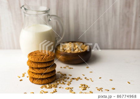 Heap oatmeal cookies and oat cereal flakes bowl on white table on milk glass jug background Heap oatmeal cookies and oat cereal flakes bowl on white table on milk glass jug background 86774968