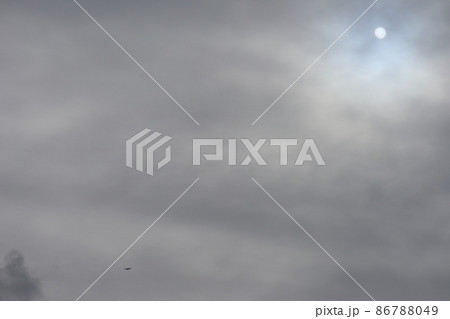 灰色雲の薄くなった青空の部分に輪郭のはっきりした太陽と小さく飛ぶ飛行機のある雲の多い冬空の風景 86788049
