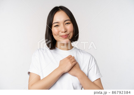 Portrait of carig young asian woman holding hands on heart, gazing with care and love, touched by something, feel flattered, standing over white background 86792384