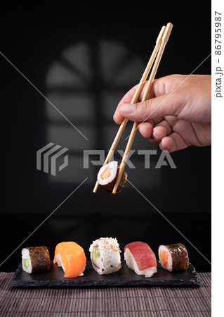 Hand picking sushi variety with chopsticks on dark background. Vertical format. Hand picking sushi variety with chopsticks on dark background. Vertical format. 86795987