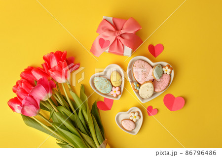 Sweets, pastry, gingerbread cookies for Easter table. Easter eggs heart shaped decor plate, pink tulips on yellow background top view copy space, spring seasonal holiday banner for site, flyer, coupon 86796486
