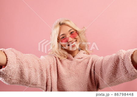 Happy blogger lady making selfie, wearing colorful glasses and hoodie, posing and smiling to camera, pink background 86798448