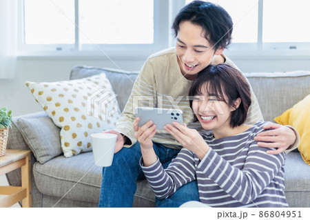 ソファーでスマホを見ながらくつろぐ若い夫婦 86804951
