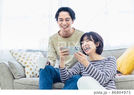 ソファーでスマホを見ながらくつろぐ若い夫婦 86804973