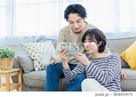 ソファーでスマホを見ながらくつろぐ若い夫婦 86804974