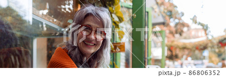 Positive mature woman with glasses and laptop holds cup ositting near cafe 86806352