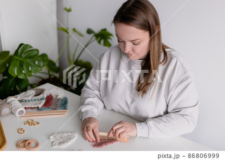 Woman working out last details of an almost-finished macrame piece, combing ropes inside a home room. Close up. Natural cotton threads and wooden beads. Female hobby. Wall hanging decor. 86806999
