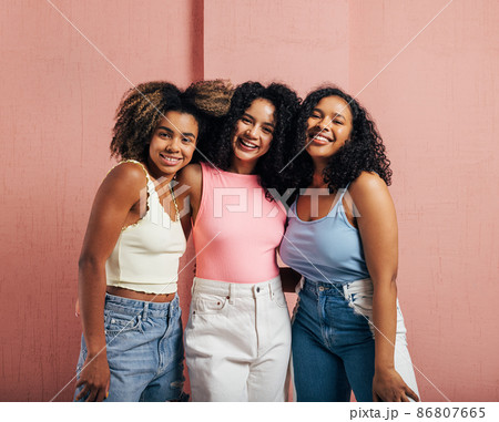 Three cheerful women with curly hair having fun together while standing at a pink wall Three cheerful women with curly hair having fun together while standing at a pink wall 86807665