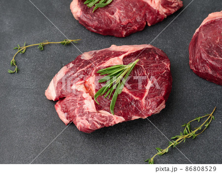 raw piece of beef ribeye with rosemary, thyme on a black table, top view 86808529