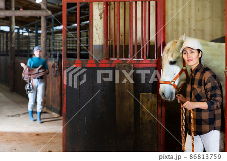 Asian female horse breeder leading white racehorse by bridle out of stall door 86813759