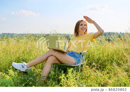 Summer vacation in wild meadow, adult woman resting on folding chair for camping using laptop 86820802