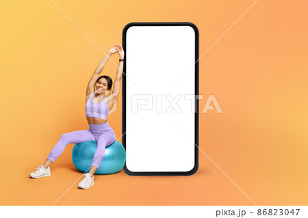 Fitness app. Athletic black lady training on fitness ball near big cellphone with blank screen, mockup Fitness app. Athletic black lady training on fitness ball near big cellphone with blank screen, mockup 86823047