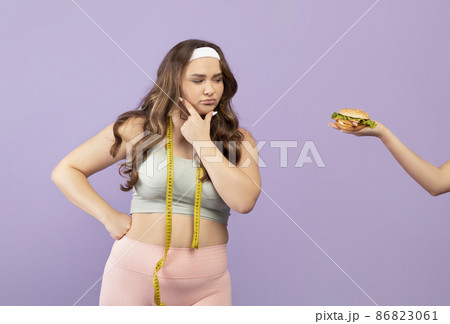 Pensive millennial caucasian plus size lady in sportswear with measuring tape looks at hand with burger 86823061
