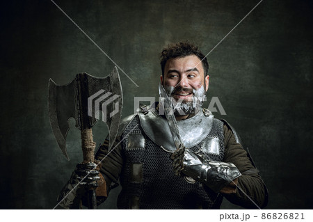 Comic portrait of medieval warrior or knight with dirty wounded face shaving with an ax isolated over dark vintage background. Comparison of eras, history, beauty 86826821