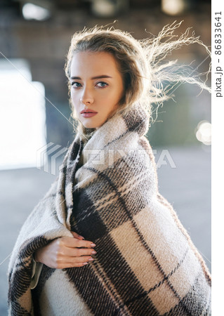 Beautiful young woman portrait wrapped in wool blanket looking to camera on windy sunny day on urban street background Beautiful young woman portrait wrapped in wool blanket looking to camera on windy sunny day on urban street background 86833641