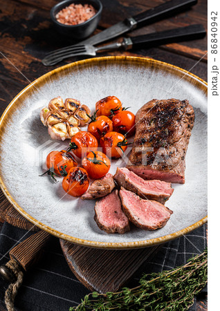 Roasted Lamb tenderloin meat in plate with grilled tomato and garlic, mutton sirloin fillet steak. Dark background. Top view 86840942