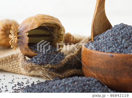 Low angle close-up of broken dry poppy seed pod with ripe poppies. White wooden table with wooden bowl full of poppy seeds. 86842119