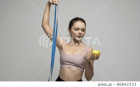 Confused attractive sexy brunette women in sport outfit standing with a measuring tape and holding a apple on a white background. Beauty and health concept. Healthy eating and diet. Confused attractive sexy brunette women in sport outfit standing with a measuring tape and holding a apple on a white background. Beauty and health concept. Healthy eating and diet. 86842523