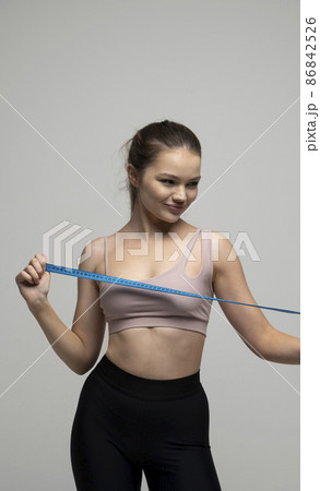 Happy attractive brunette woman with a perfect sexy body in beige top sportswear holding a blue measuring tape. Weight loss and diet concepts. Health care and healthy nutrition. Perfect slim body. 86842526