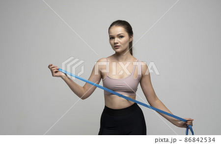Weight loss and diet concepts. Attractive sporty slim brunette female in sportswear holding a blue measuring tape and looking in a camera. Health care and healthy nutrition. 86842534