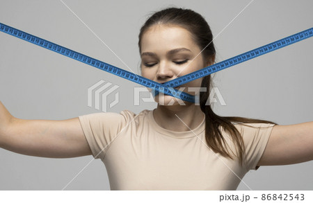 Young attractive beautiful brunette woman in a beige outfit holding measuring tape around her mouth on white background. Healthy lifestyle and diet concept. 86842543