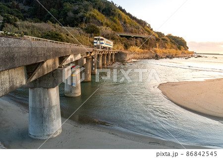 日向の南部の太平洋に昇るオレンジ色の朝日をうけて岩場の海岸線を走る日南線白いキハ40 86842545