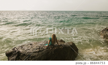 Woman sitting on rock of sea reef stone, stormy cloudy ocean. Blue swimsuit dress tunic. Concept resort coastline tourism summer holidays 86844753