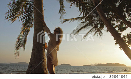Woman drinking pineapple cocktail Pina Colada near palm tree, in white shirt, sunglasses. Concept rest tropical sea traveling tourism summer holidays 86844845