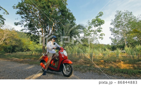Man on red motorbike in white clothes drive on forest road trail trip. One men caucasian tourist go on scooter, nearby tropical palm tree. Man on red motorbike in white clothes drive on forest road trail trip. One men caucasian tourist go on scooter, nearby tropical palm tree. 86844883
