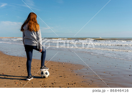 Spanish pre-adolescent girl looking at the horizon on the shore of Burriana beach stepping on her soccer ball with one foot. Concept of illusion and hope to achieve your dreams. 86846416