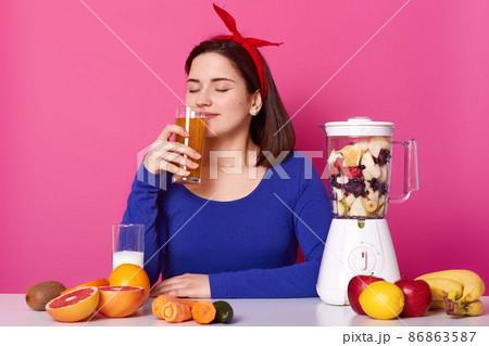 Close up portrait of pleasant charming young lady smelling fruit smoothie with closed eyes, enjoys smell, looks happy being on diet. Positive model sits isolated over bright pink background. 86863587