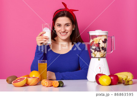 Smiling European woman holds glass of milk, prepares homemade healthy fruit smoothie, surrounded with grapefruit, apples, bananas, carrot, kiwi, sits at kitchen table against pink background Smiling European woman holds glass of milk, prepares homemade healthy fruit smoothie, surrounded with grapefruit, apples, bananas, carrot, kiwi, sits at kitchen table against pink background 86863595