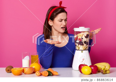 Puzzled woman cannot understand why food processor doesnt work, lgestures with both hands, fails to switch it on, wears red headband and blue jumper, isolated over pink wall. How to make smoothie? 86863614