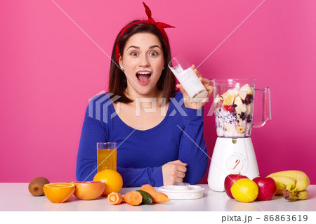 Healthy food concept. Close up of a young woman uses berries and bananas for making smoothie. Astonished lady againest pink studio wall drinks milk while preparing coktail. Healthy lifestyle concept. Healthy food concept. Close up of a young woman uses berries and bananas for making smoothie. Astonished lady againest pink studio wall drinks milk while preparing coktail. Healthy lifestyle concept. 86863619
