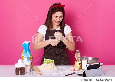 Happy housewife wears kitchen apron dirty with flour, white t shirt, red headband, kneads dough while having videocall with her husband. Studio picture of smiling baker isolated over pink background. Happy housewife wears kitchen apron dirty with flour, white t shirt, red headband, kneads dough while having videocall with her husband. Studio picture of smiling baker isolated over pink background. 86863924