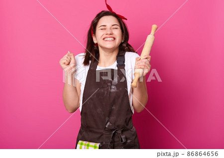 Baker woman holds baking rolling pin, wears brown apron, white t shirt. Young girl bends elbows and shouts yes with closed eyes, female maskes cake first time. Cook isolated on pink background. 86864656