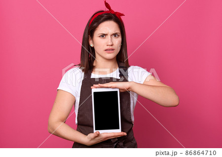 Close up portrait of woman wearing brown apron and white t shirt, showing blank empty screen with copy space for advertisment or promotion. Caucasian baker poses with her gadget, being in bad mood. Close up portrait of woman wearing brown apron and white t shirt, showing blank empty screen with copy space for advertisment or promotion. Caucasian baker poses with her gadget, being in bad mood. 86864710