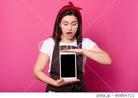 Surprised young woman holding digital tablet with blank screen, looks down, keeps mouth opened, has astonished facial expressions, sees something shocking, free space for promotion, pink background. 86864711