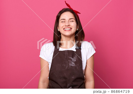 Indoor shot of beautiful magnetic cute young woman standing isolated over pink background in studio, closing eyes, smiling sincerely, wearing brown dirty apron, white t shirt and red headband. 86864719