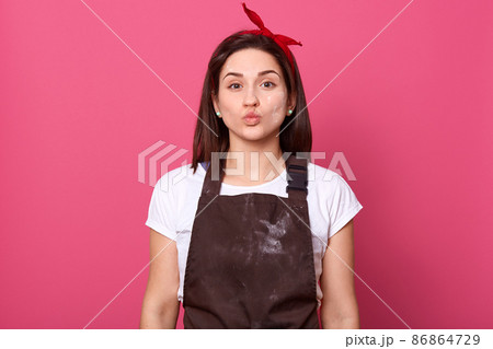 Horizontal portrait of good looking attractive sweet female pulling her lips, looking directly at camera, having dirty brown apron, wearing red headband and white t shirt, being hard working. 86864729