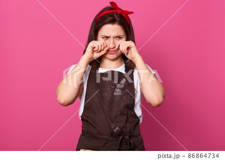 Disappointed upset housewife touching her face with hands, wiping tears out, crying, wearing brown apron, white t shirt and red headband, being in bad mood, tired of household chores. Routine concept, 86864734