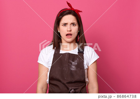 Portrait of impressed attractive female wearing brown dirty apron, white t shirt, having shocked facial expression, opening mouth widely with indignation, isolated over pink background in studio. 86864738
