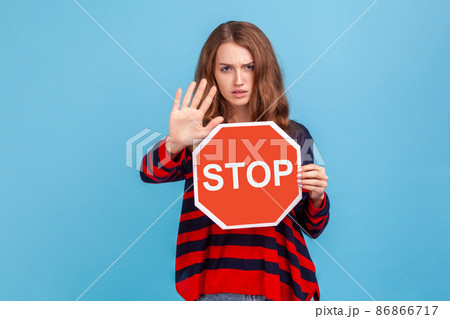Strict woman wearing striped casual style sweater, looking angrily, showing ban gesture, holding stop sign as warning of ban, forbidden access. Indoor studio shot isolated on blue background. 86866717