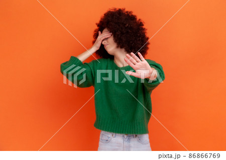 Depressed woman with Afro hairstyle wearing green casual style sweater covering eyes with palm and showing stop ban gesture with palm. Indoor studio shot isolated on orange background. 86866769