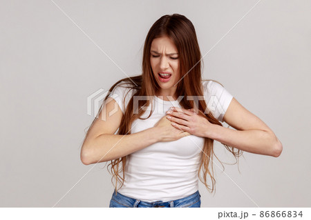 Portrait of woman clutching chest, suffering sudden ache, painful cramps, risk of infarction, cardiac problems, wearing white T-shirt. Indoor studio shot isolated on gray background. Portrait of woman clutching chest, suffering sudden ache, painful cramps, risk of infarction, cardiac problems, wearing white T-shirt. Indoor studio shot isolated on gray background. 86866834