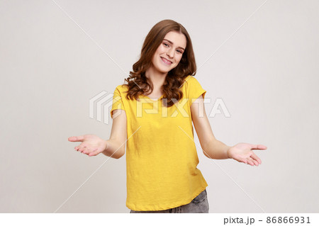 Please, take for free. Friendly pretty young woman in yellow T-shirt welcoming with wide open arms and smiling kindly, looking smiling at camera. Indoor studio shot isolated on gray background. 86866931